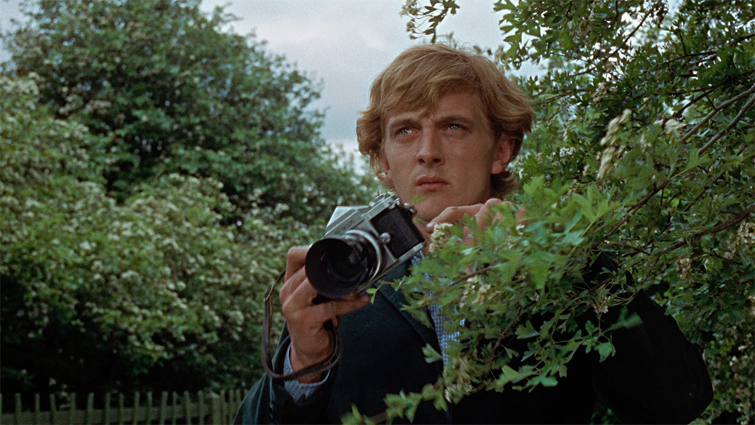A man with shaggy hair stands behind a branch and he holds a camera. He is staring with a look of suspicion into the distance