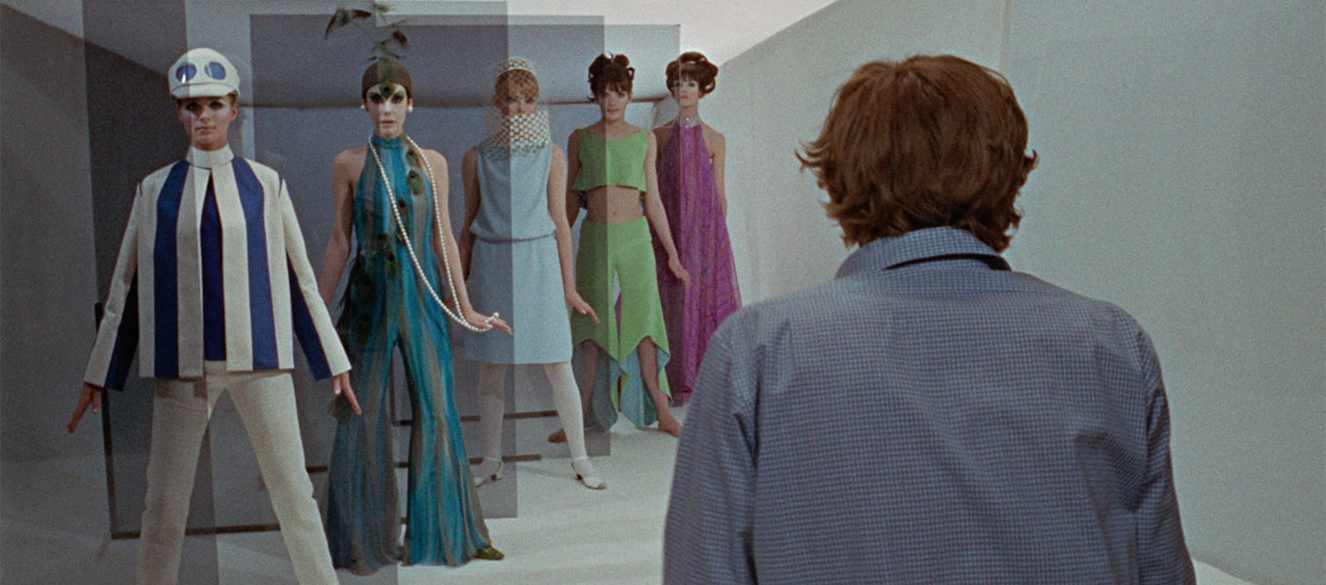 A man in a blue shirt and shaggy hair stands with his back to us. He is looking forward at a row of five fashion models in stylish, mid-1960s mod attire