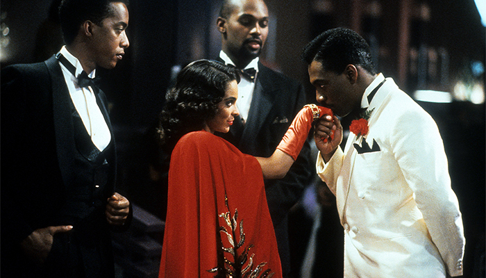 Eddie Murphy, dressed in dapper 1930s style in a white suit with a red rose in the lapel kisses the gloved hand a woman in a red dress