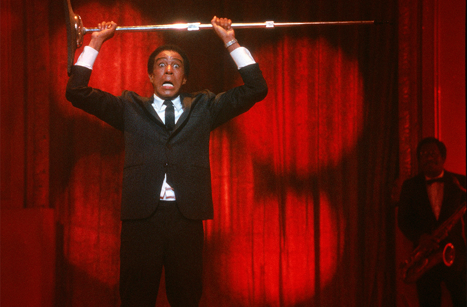 Richard Pryor stands on stage in front of a red curtain and holds a microphone stand above his head. His mouth is open in mock surprise