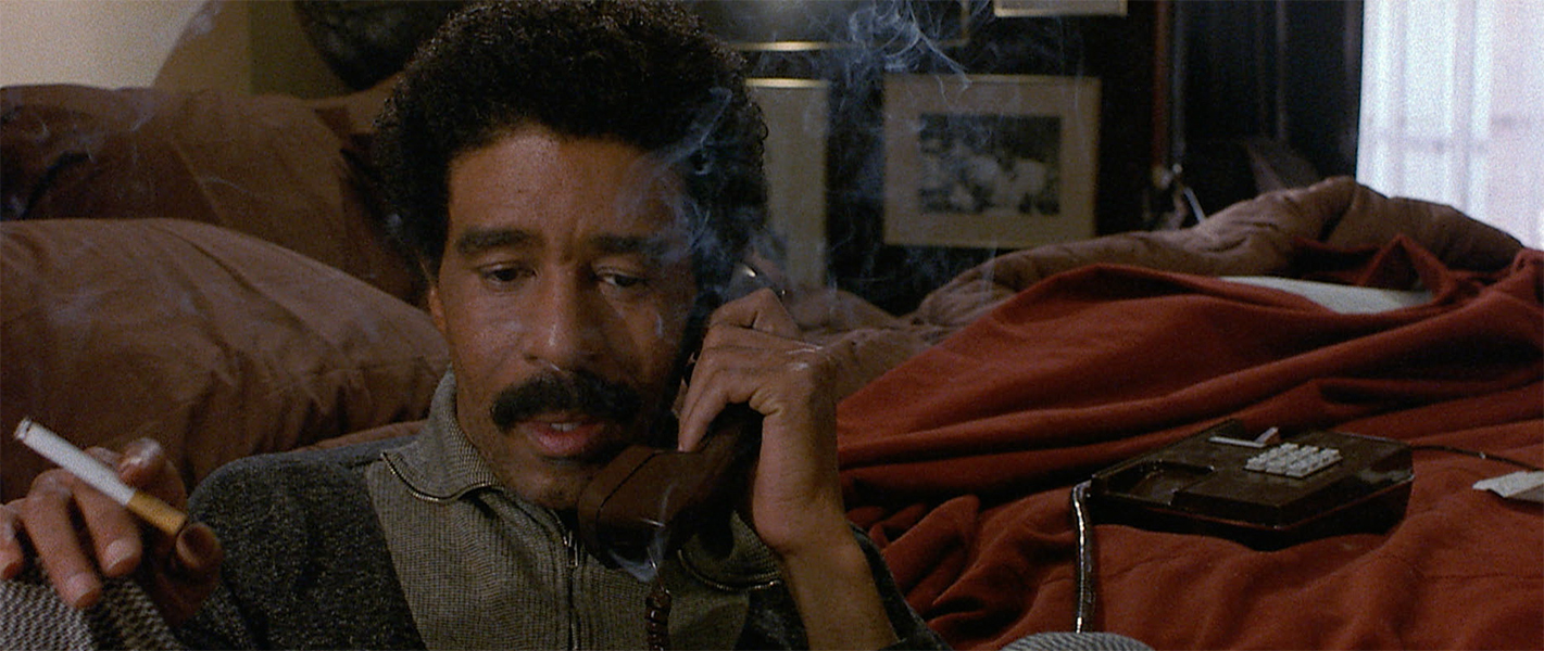 Richard Pryor is seated and on the phone while smoking a cigarette