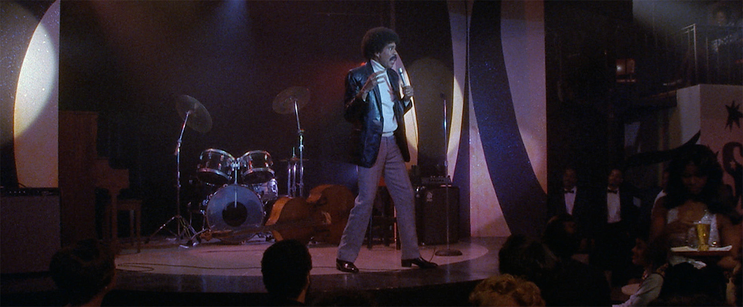 Richard Pryor on stage in a dark, smoky club. He wears a leather jacket and brown pants.