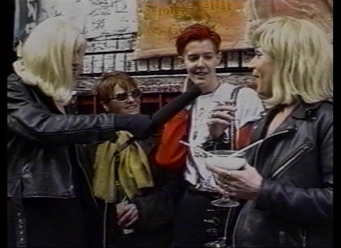 A color photo of Glenn Belvario interviewing attendees to a Riot Grrls conference in 1993. Glenn and his cohost are dressed in blonde wigs and leather biker jackets.