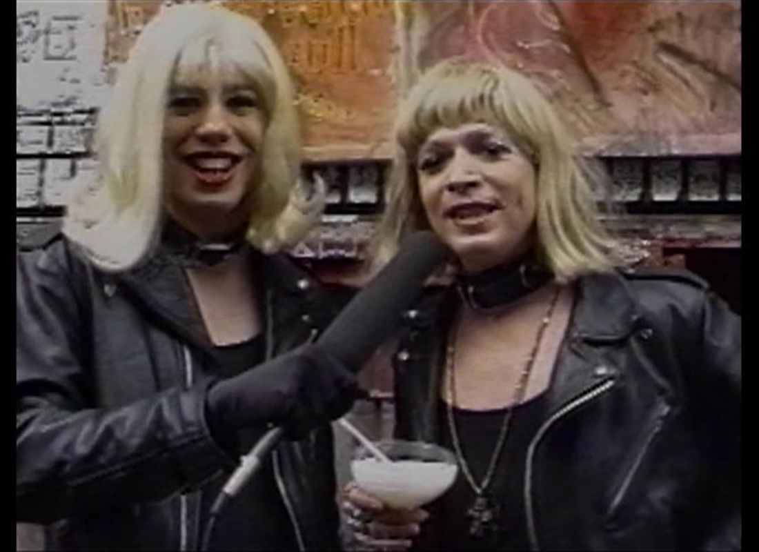 A color photo of Glenn Belvario holding a microphone to cohost Fonda LaBruce; both are wearing blonde wigs and leather biker jackets.