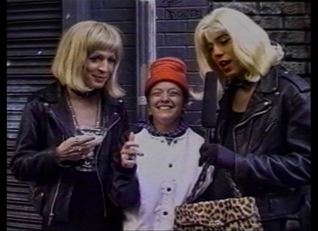A color photo of Glenn Belvario and cohost Fonda LaBruce wearing blonde wigs and leather biker jackets interviewing a person in a white coat and red hat.