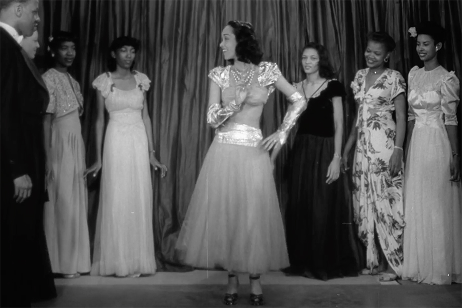 A group of six women stand on a stage, all dressed to the nines in gowns and evening wear. A man stands off to the side looking at them. In the center stands one woman looking over at the men watching them