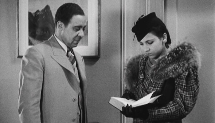 A man and woman stand in a doorway. The woman is in a fur coat and is looking down at a book in her hands