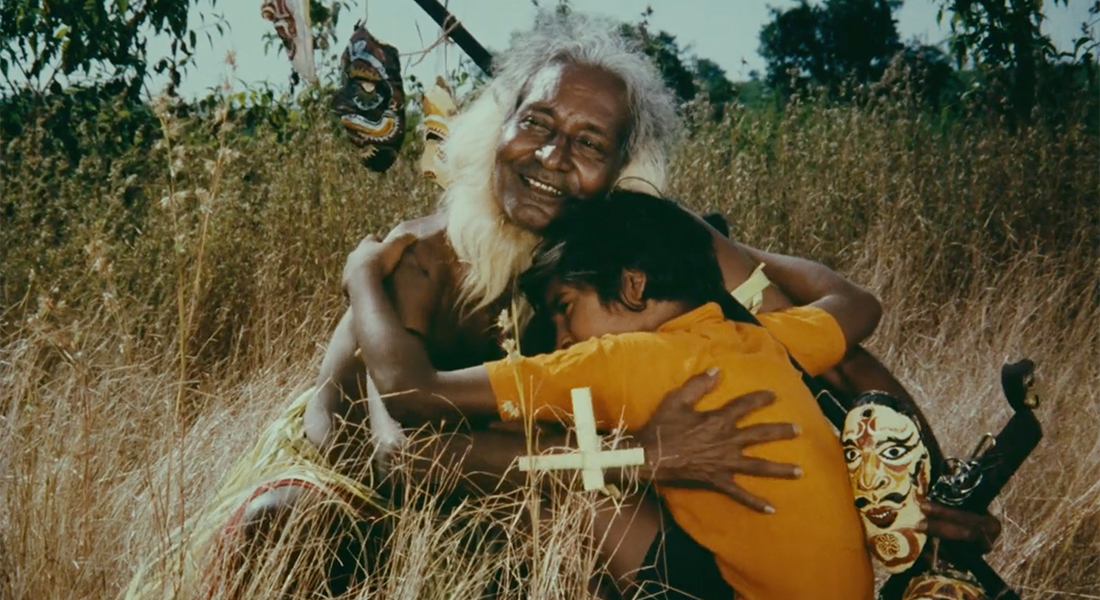 An elder with white hair smiles and holds a young child in a yellow shirt. A cross sits in front of them as they both sit in a field