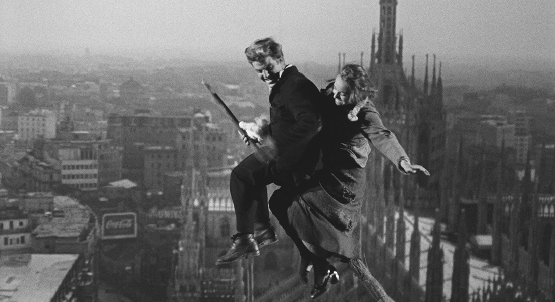 An image of a man and a woman on a broomstick flying above the tops of buildings in a city