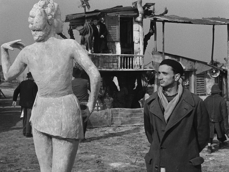 A man in a heavy overcoat stars at the statue of a woman. Behind him is a shabby building, falling apart 