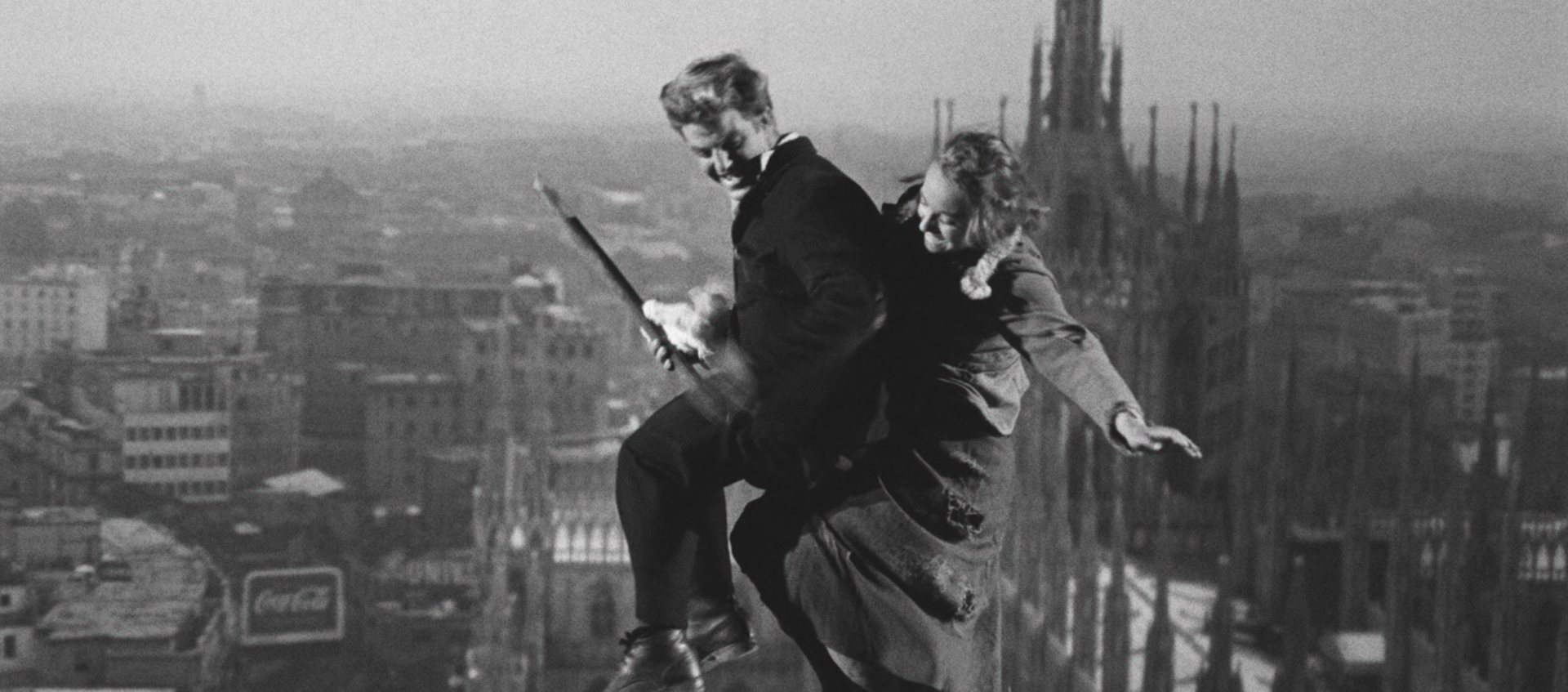 An image of a man and a woman on a broomstick flying above the tops of buildings in a city