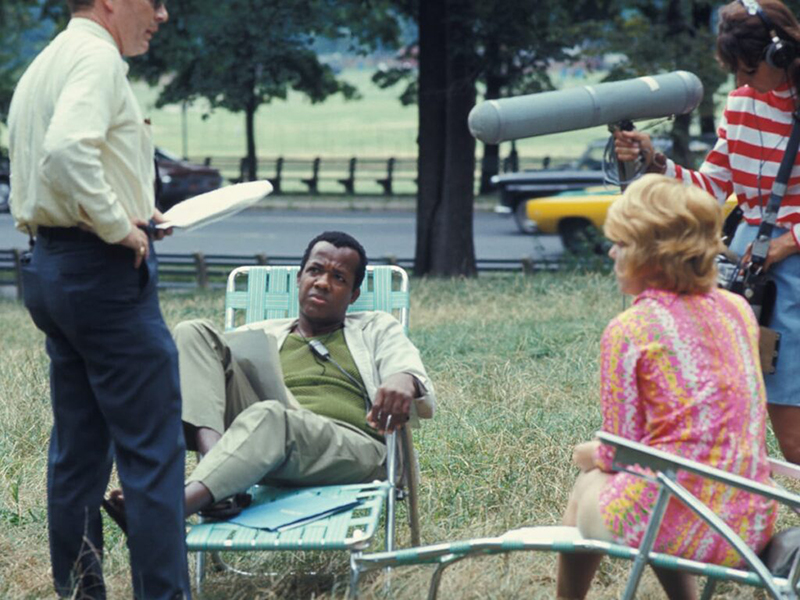 A group of four people sit in a grassy area. One man in brown sits in a lawn chair, a woman in a pink skirt and short blonde hair sits in a lawn chair. To the man's left stands another man holding a script. To the woman's right stands a man in a red and white striped shirt holding a boom microphone.