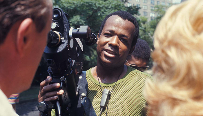 Filmmaker William Greaves stands in front of a man and a woman. He is holding a film camera.
