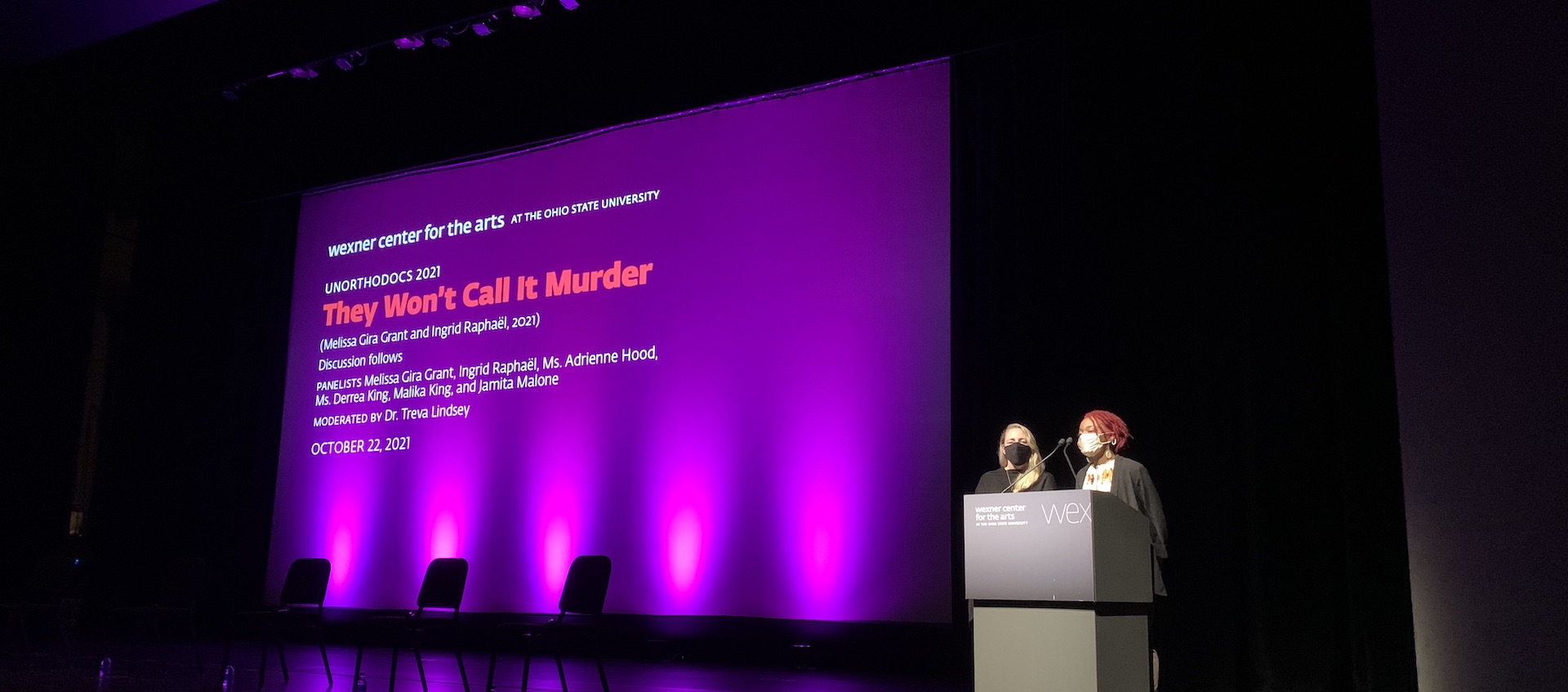 Melissa Gira Grant and Ingrid Raphael stand on the stage of Mershon Auditorium. Lighting casts a purple glow up to a movie screen to the right, which holds a graphic with the title of their film, They Won't Call it Murder