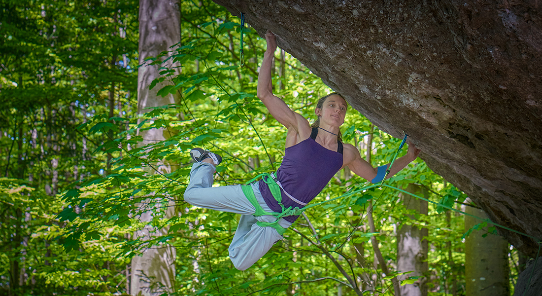 A person in mountain climbing gear--a tank top and gray pants is in a harness while dangling off the side of a rock