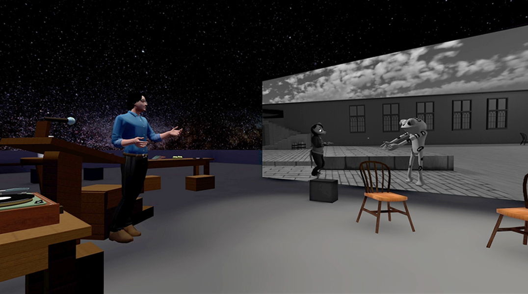 In a computer-animated still a person in a blue shirt seems to speak on a virtual stage, gesturing to a black-and-white animation of a frog interacting with a humanoid rodent