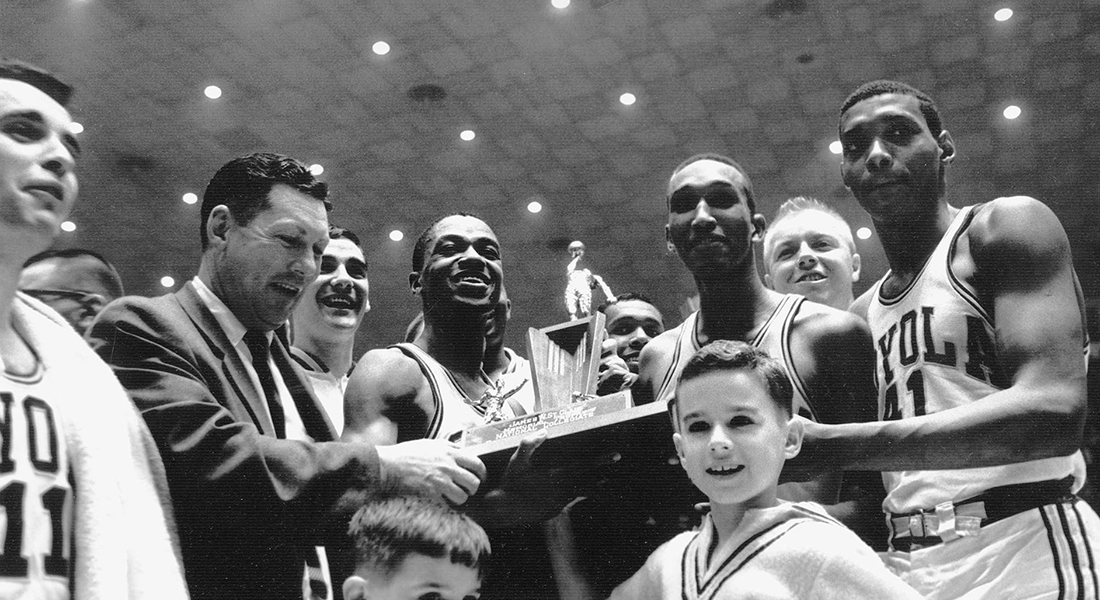 A basketball team gathers around one another as three players hold a trophy