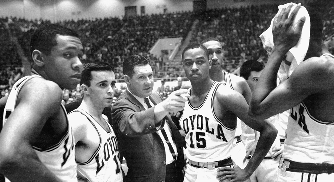 Six basketball players in Loyola jerseys on the court flank a coach who is wearing a suit. The coach points to the basketball court and the players look in the same direction.