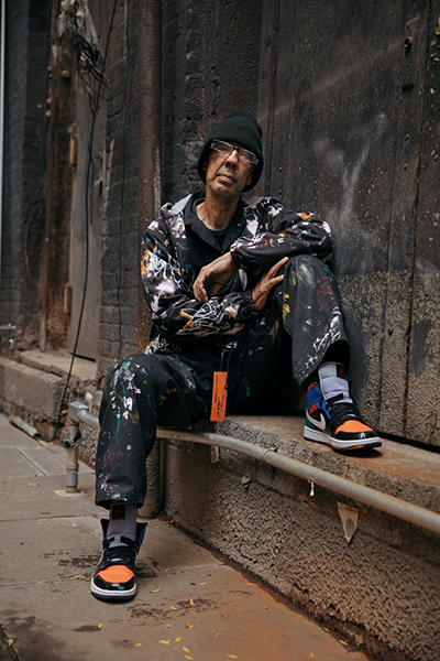 Futura2000 in a black wool cap wears paint-splatterd clothes, brightly colored sneakers, and sits with one leg propped on a cement bench