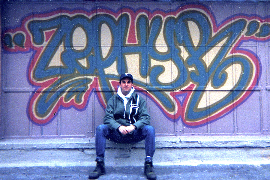 Zephry sits in front of a brightly colored spray painted wall with his name spray painted in bright, vivid colors