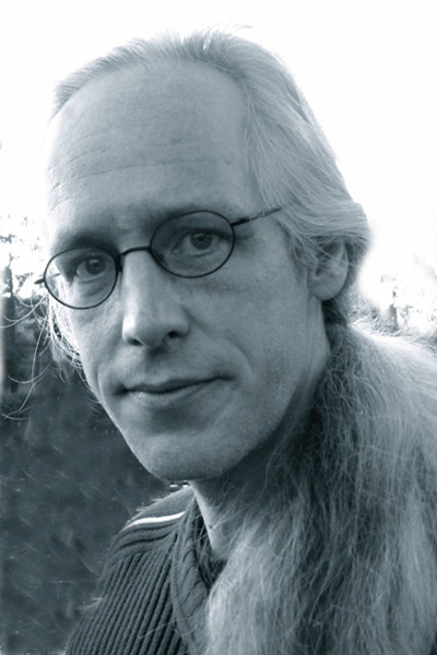 Carlo McCormick with long gray hair in a ponytail that sits on their shoulder. he wears thin wire-rimmed glasses