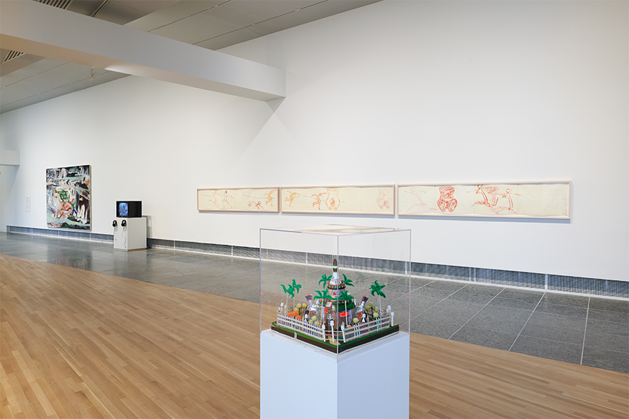 A photograph of a museum gallery featuring a large painting, a video monitor on a pedestal, and a long rectangular drawing hung on a wall in the background. A small sculpture sits atop a pedestal in the foreground.