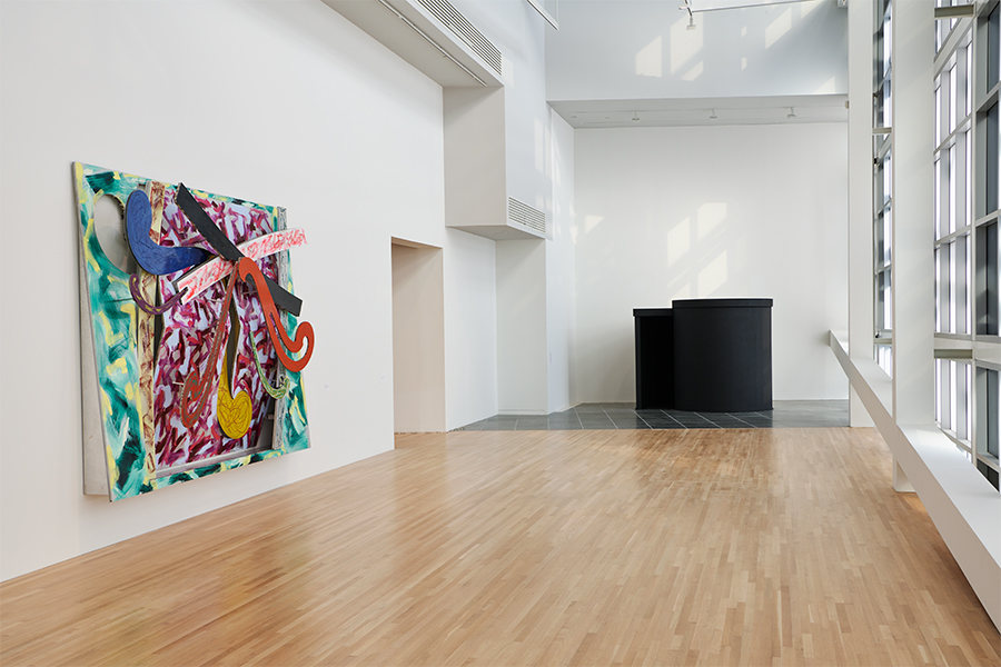 A photograph of Frank Stella's painted aluminum relief Puerto Rican Blue Pigeon (1976) and Adrian Piper's immersive installation Four Intruders Plus Alarm Systems (1980) in the exhibition To Begin, Again: A Prehistory of the Wex, 1968–89.