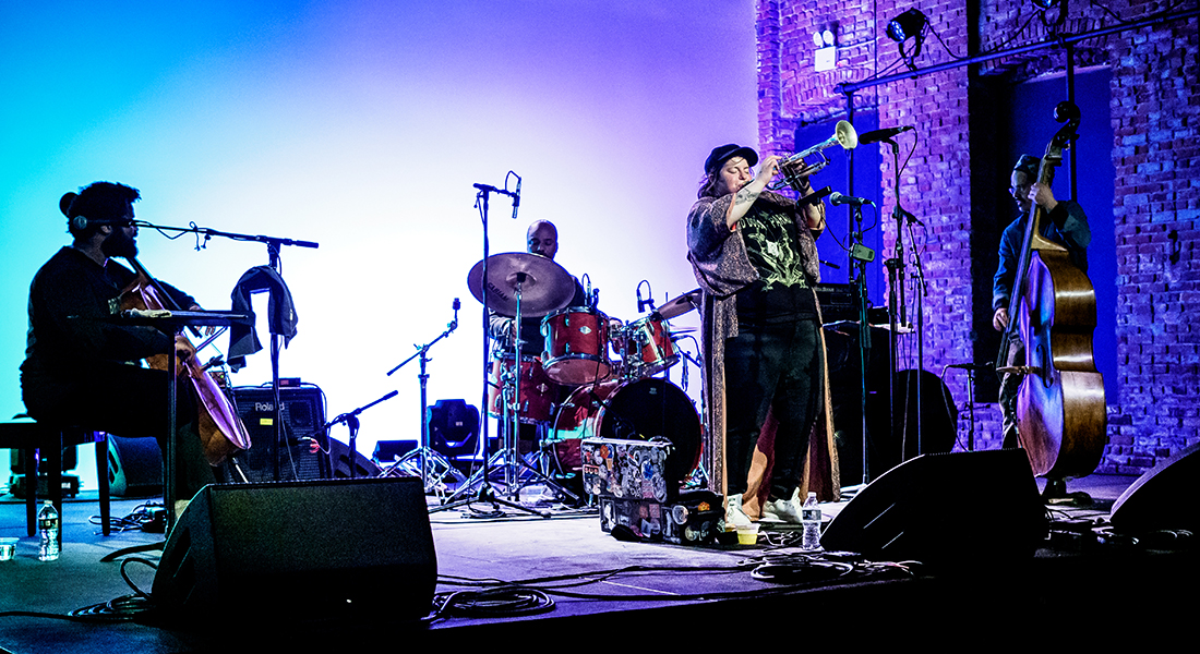 jaimie branch holds her trumpet aloft at the front of a stage with the members of FLY or DIE playing cello, drums, and upright bass around her. At the back of the stage is a white scrim lit in blues and purple; at the right of the image is an exposed brick wall.