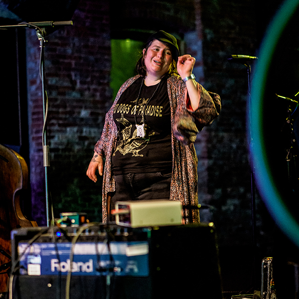 jaimie branch wearing a patterned long shirt and black concert t-shirt that reads “The Dogs of Paradise” smiles and gestures with her right hand. A brick wall is in the back of the shot and amplifiers are in the foreground.