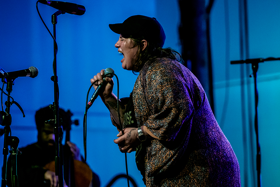 jaimie branch wearing a patterned long shirt and dark cap screams into a microphone she’s holding; she holds the cord in her other hand. In the background is a person playing cello, out of focus, a black boom mic stand; and a scrim lit in blue.