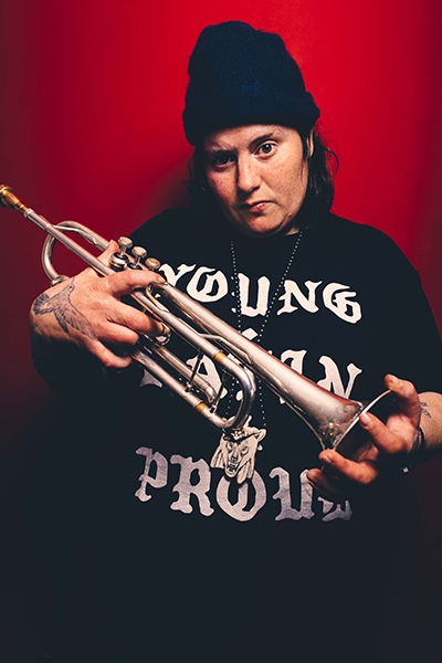 jaimie branch holds her trumpet to the camera wearing a black cap and t-shirt that reads "young, latin, proud." She is photographed against a red background.