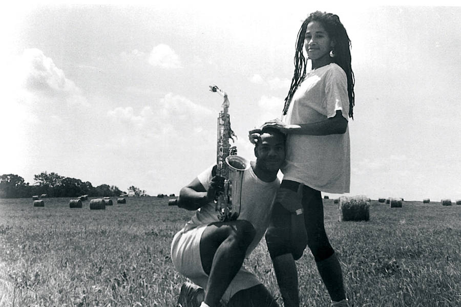 Black-and-white film still from A Powerful Thang featuring main characters Craig (left) and Yasmine (left) in a field of hay bales. Craig is kneeling with one arm around Yasmine and the other holding his saxophone. Yasmine is standing with her hands on Craig’s head. 