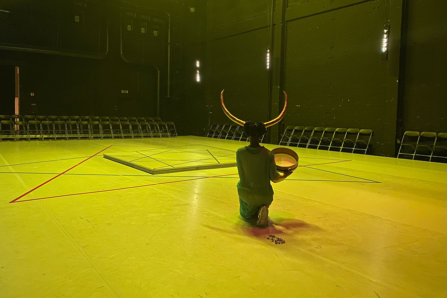 erformer Dorothée Munyaneza, who is wearing large golden horns in her dark hair and a blue transparent dress, kneels, with her head upright and back towards the camera, next to a round object on the floor in a space surrounded by chairs. The space is tinted with yellow-green light.