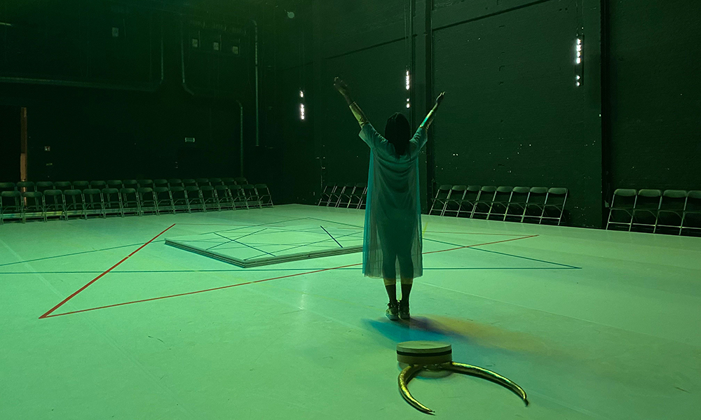 Performer Dorothée Munyaneza, who is wearing a blue transparent dress, stands with hands above her head facing the center of a stage, her back turned to the camera, with large golden horns behind her. The space is tinted with green-blue light.