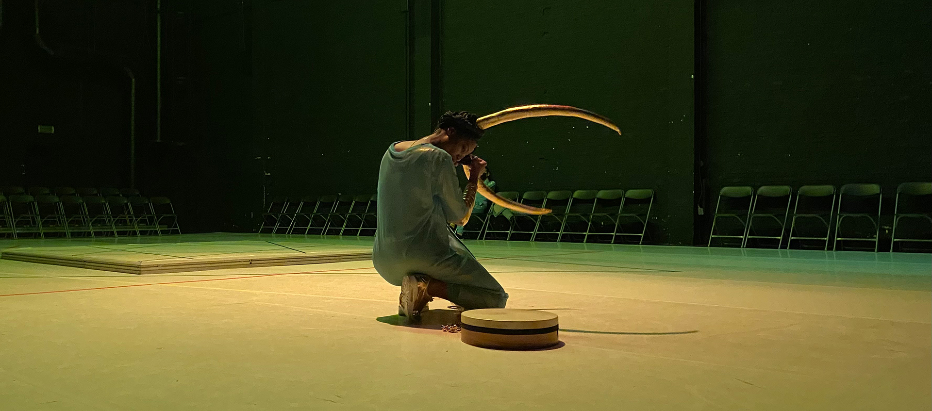 Performer Dorothée Munyaneza, who is wearing large golden horns in her dark hair and a blue transparent dress, kneels beside a round object on the floor in a space surrounded by chairs. The space is tinted with yellow-green light.