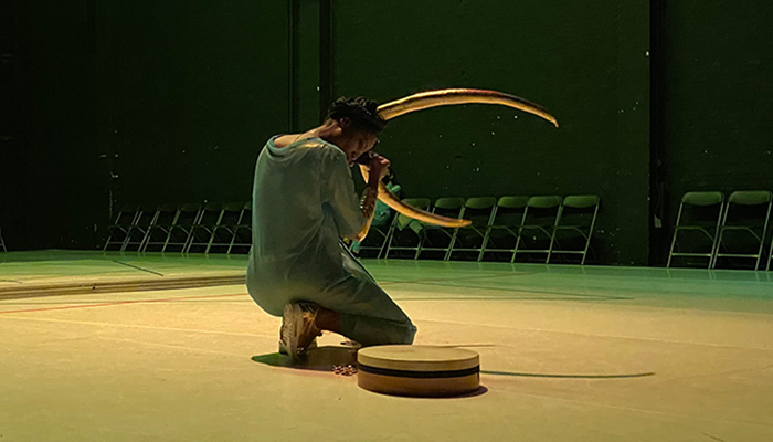 Performer Dorothée Munyaneza, who is wearing large golden horns in her dark hair and a blue transparent dress, kneels, with her head pointing towards the ground and her profile in view, beside a round object on the floor in a space surrounded by chairs. The space is tinted with yellow-green light.