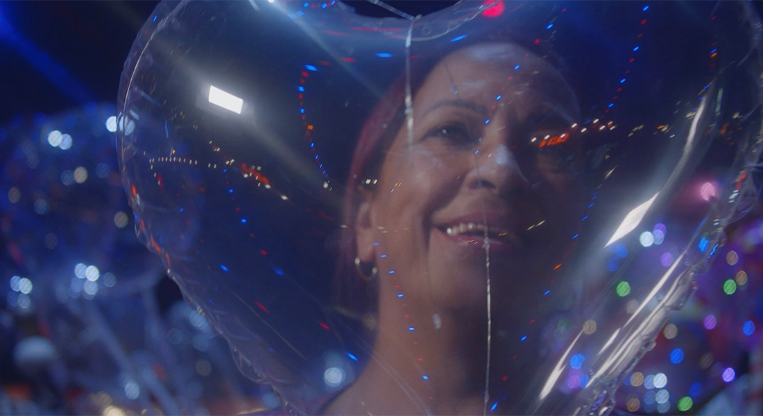 Still of a person with dyed red hair and brown skin who is smiling behind a transparent, heart-shaped balloon, with colorful lights twinkling throughout the image.