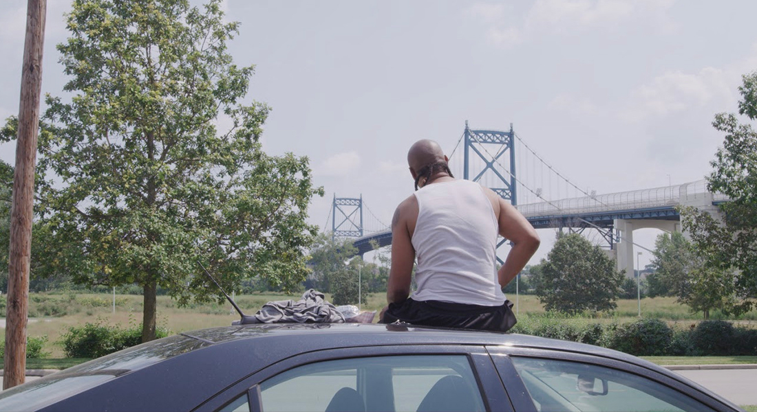 Still of a person with brown skin, a bald head, and white no-sleeve shirt who is sitting on top of a car, back to the camera, facing a bridge and greenish-yellow landscape in the background. 