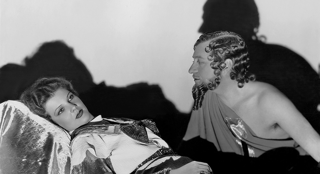 Black-and-white still from The Warrior’s Husband featuring a woman in warrior garb laying down while a man (on her right) with curly hair and a long beard looks over her.