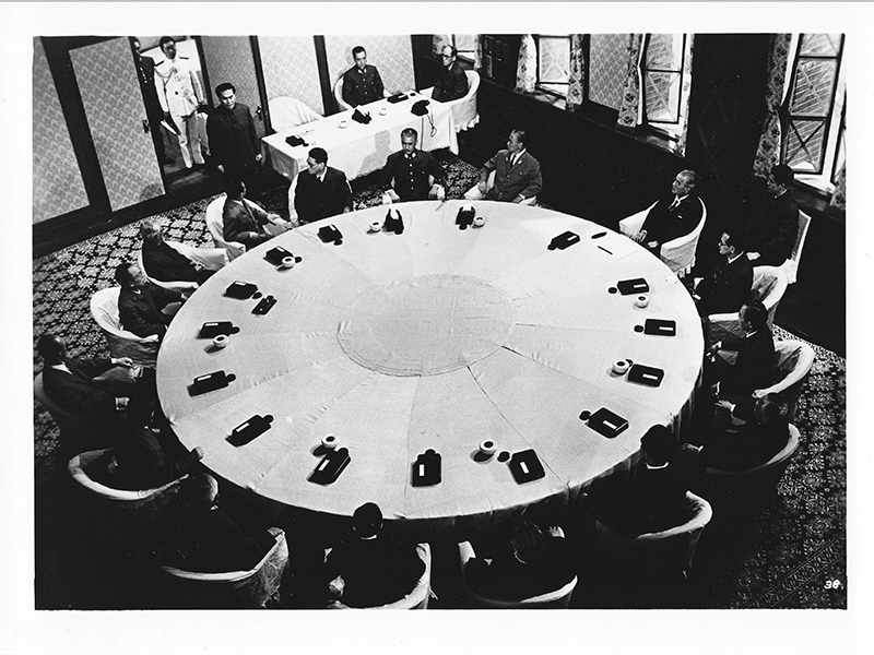 Black-and-white still of people wearing suits and seated around a large, round table.