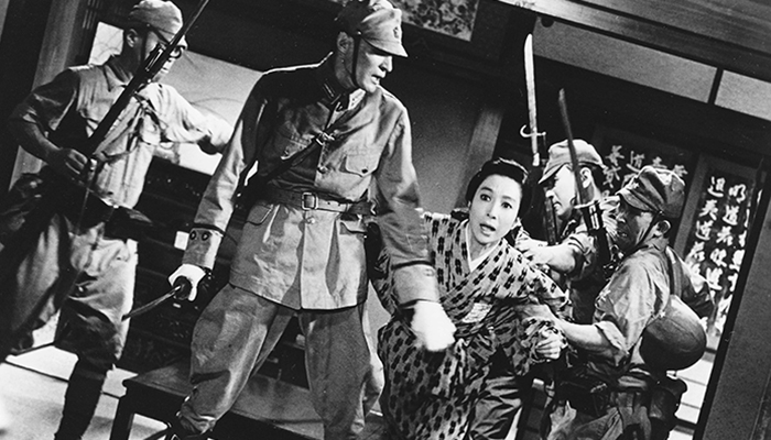 Black-and-white still of a person in a patterned kimono who is trying to flee from four soldiers with weapons.