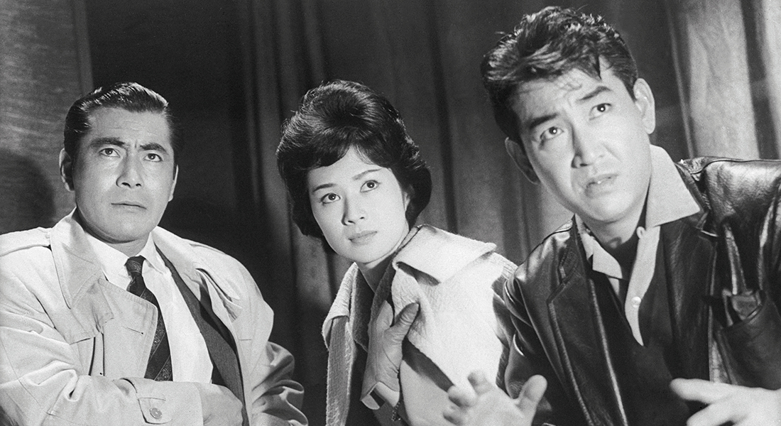 Black-and-white still of three people looking intently at something; the person on the left has short, slicked-back dark hair and is wearing a suit, tie, and trench coat; the person in the middle has dark, short, voluminous hair and is wearing gloves and a light-colored jacket; the person on the right has short, dark hair and is wearing a black leather jacket. 