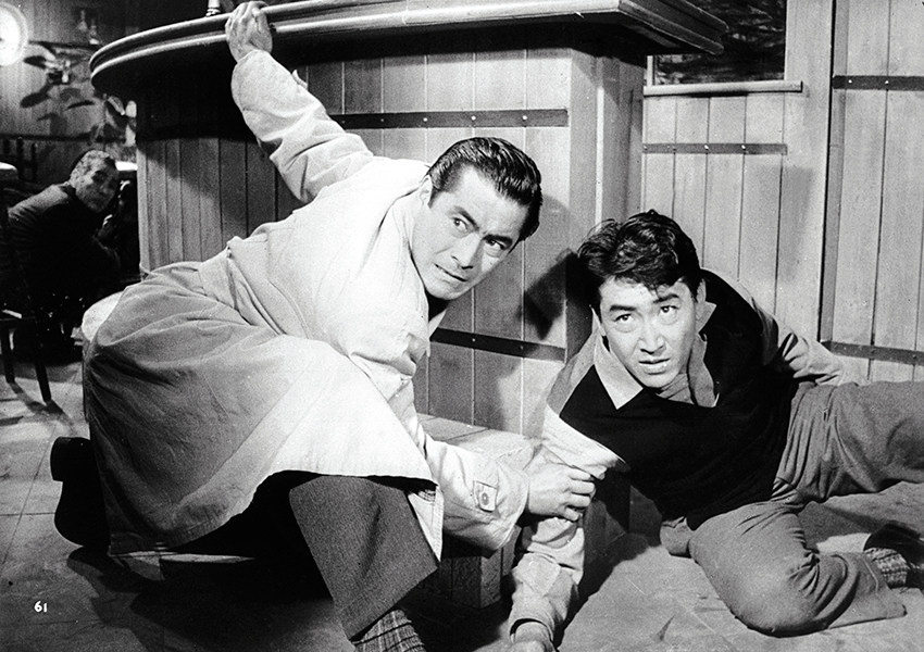 Black-and-white still of two people crouched behind a bar; the person on the left has one hand gripping the bar counter and the other gripping the arm of the person on the ground next to them.