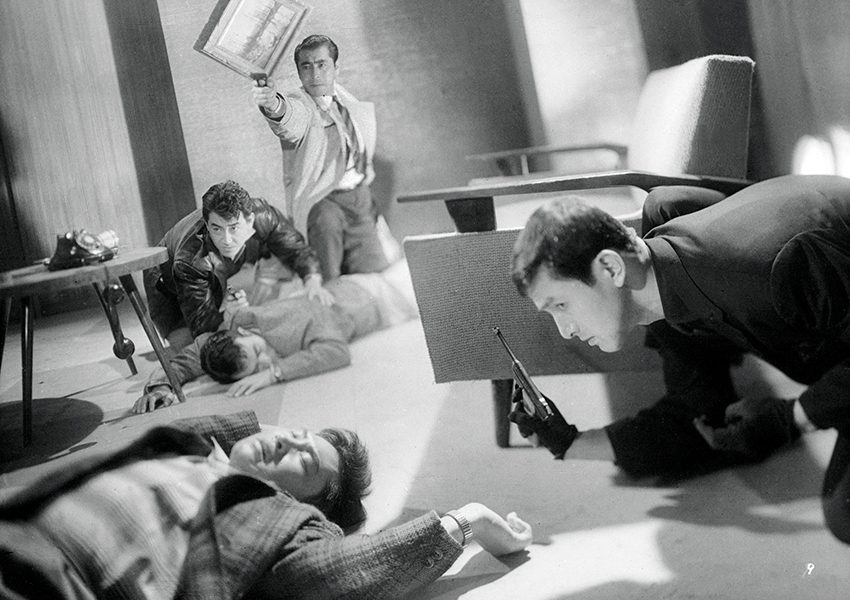 Black-and-white still of a gunfight where three people are crouched on the ground with guns pointed in front of them, along with two unconscious people on the floor in what appears to be a living room. 