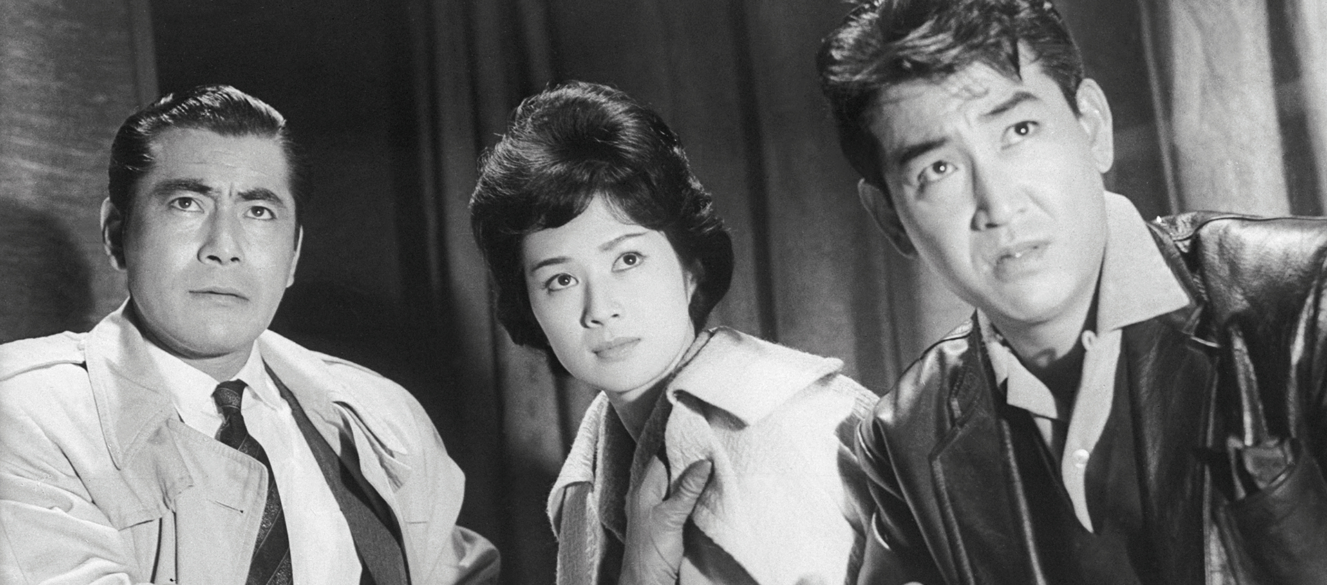  Black-and-white still of three people looking intently at something; the person on the left has short, slicked-back dark hair and is wearing a suit, tie, and trench coat; the person in the middle has dark, short, voluminous hair and is wearing gloves and a light-colored jacket; the person on the right has short, dark hair and is wearing a black leather jacket. 