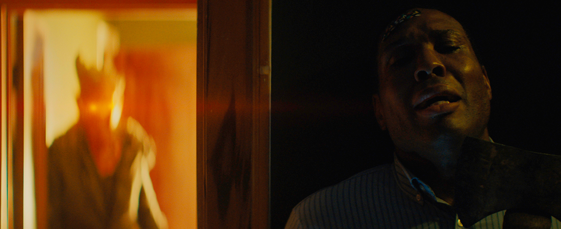 Still of a person hiding behind a door, looking terrified, with a bandaid on their head. To their left in the background is a blurred figure wearing a mask and lit in red light.