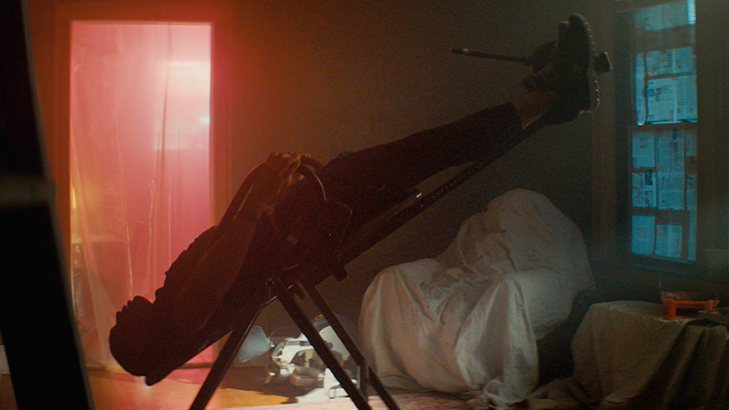 Still of a person on a stretcher with head angled toward the floor and feet in the air; the door of the room is draped in a see-through material lit up by red light in the background.