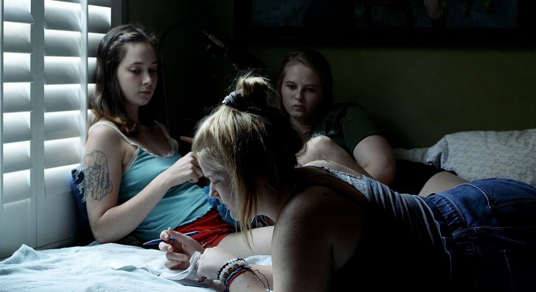 Still of three young people on a bed. The person in the foreground is lying on their stomach with a pen in one hand and a notepad in the other