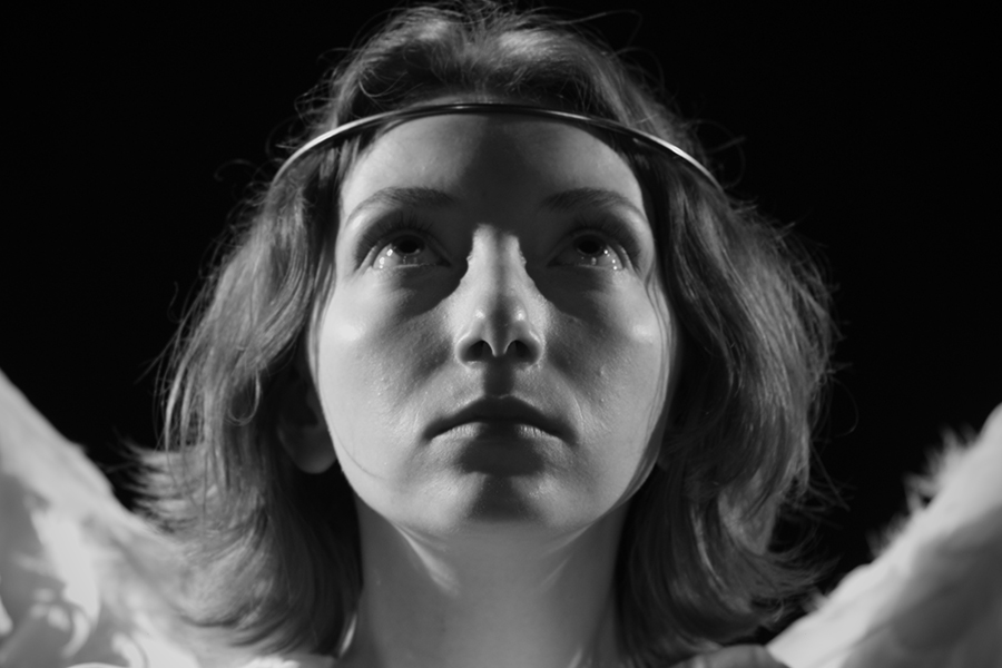 Black-and-white still of a young angel with a metal halo around their head and the tops of their white wings behind them