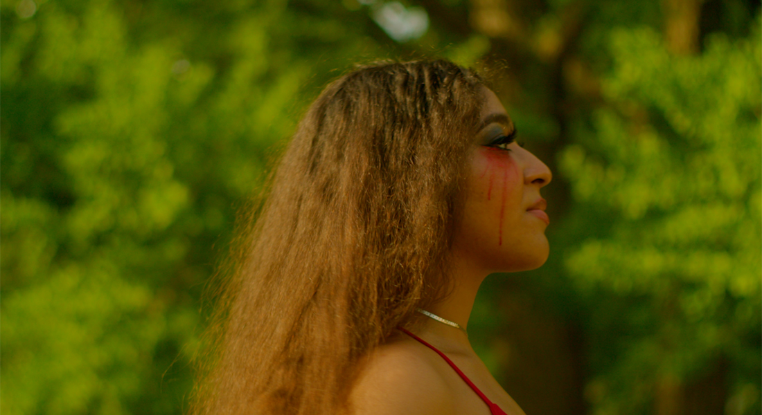 Still of the profile of a person with long brown hair and red makeup streaming down their face; blurred trees are in the background. 
